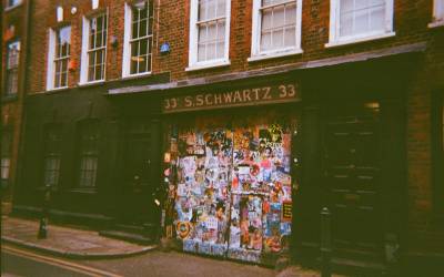grainy photograph of a disused shop front in an Edwardian terrace. The shop sign reads "33 S. Schwartz 33" and the front of the shop is plastered with posters and stickers.