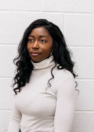 young black woman with long dark hair and white t-shirt stands against white wall and looks slightly off camera