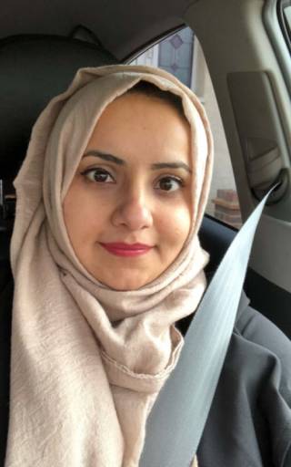 Portrait of women sat in a car, wearing seatbelt. She is wearing a beige hijab and bright lipstick