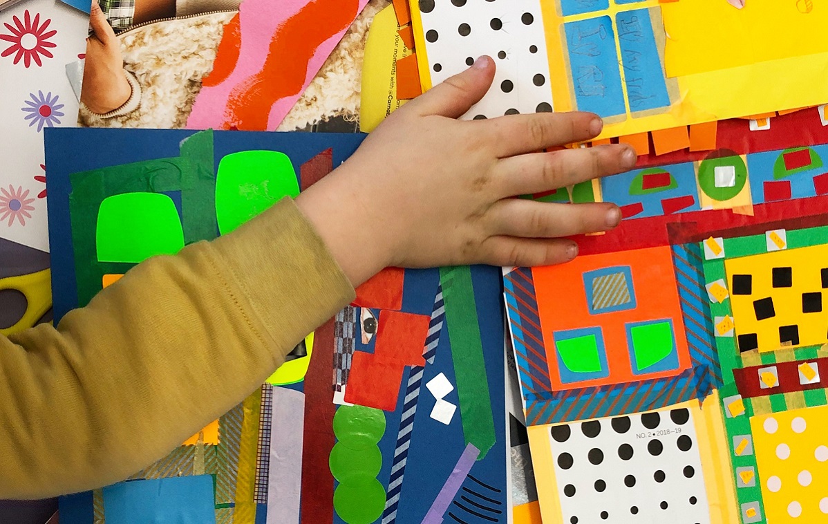 A child's hand is shown from above, reaching across a table of colourful craft materials