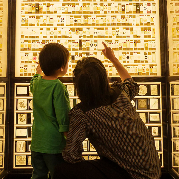 A woman crouches down next to a toddler in front of a wall of backlit microscope slides