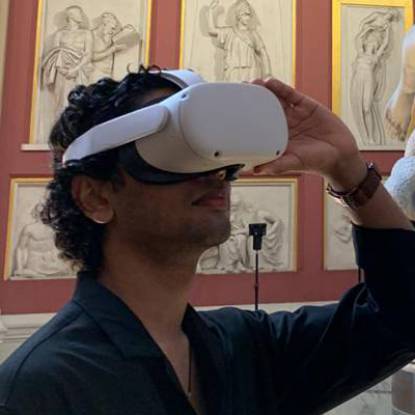 A man with dark hair wears a VR headset in a gallery space with friezes behind him on the wall