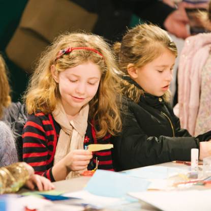 Two young girls sit side by side at a table covered in craft materials including paper, scissors and glue
