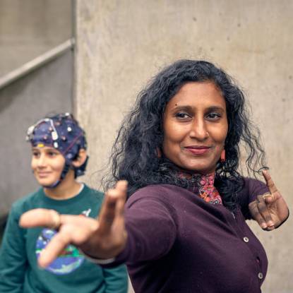 A woman reaches her arm out towards the camera as if dancing. Behind her is a young boy wearing a brain-monitoring headset.