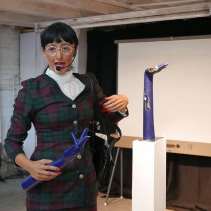 A woman with dark hair wearing a tartan dress stands in front of a projector screen, speaking into a headset and holding a blue model hand