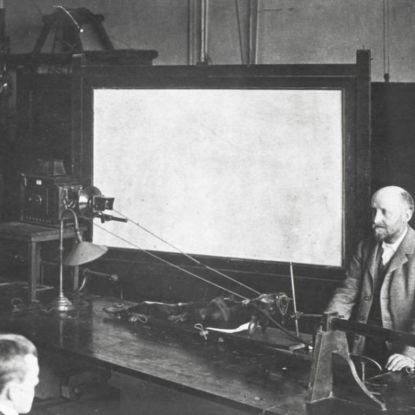 Historical black and white photograph of a man operating a piece of scientific equipment arranged across a workbench