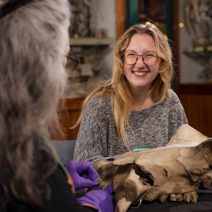 A young woman with long hair sits at a table in the Grant Museum in front of a large animal skull specimen, smiling towards an older woman who is facing away from the camera