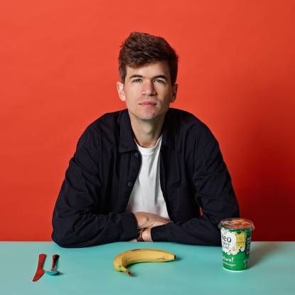 Picture of comedian Ivo Graham sitting at a table with his arms folded. On the table in front of him is a knife and spoon, a banana, and a pot of yoghurt