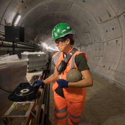 A woman with short dark hair wearing hi-vis orange and a green hard hat touches a turntable that is set up on a pallet inside an Underground tunnel