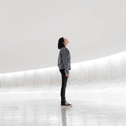 A figure in dark clothes stands looking up in a wide white space