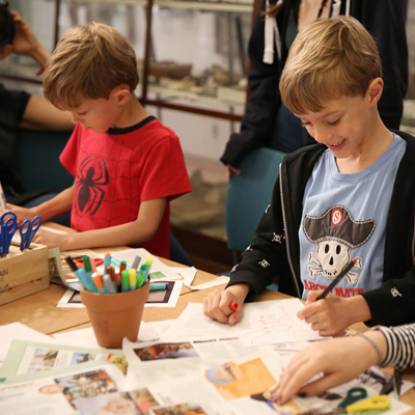 Two young boys in red and blue T-shirts work side by side at a crafting table using paper, pens and scissors