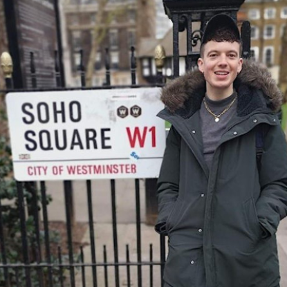 A young man in a black coat and cap stands smiling next to railings and a sign reading Soho Square