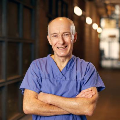 Professor Martin Birchall in scrubs with arms folded smiles at the camera. There is a long corridor behind him.