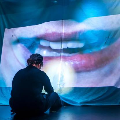 A figure in dark clothing sits with their back to us on the floor. In front of them is a large sheet hung from the ceiling with a close-up image of a mouth projected onto it.