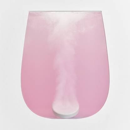 A glass-shaped transparent vessel filled with pink liquid with a white tablet dissolving at the bottom