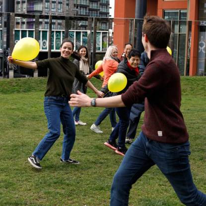 A group of people run smiling across a field holding yellow balloons