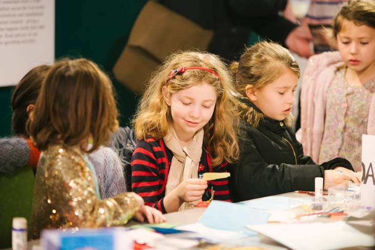 A group of young girls sit at a table covered in craft materials including paper, scissors and glue