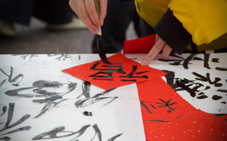 A hand brushes Chinese calligraphy onto red and white paper using a paintbrush and black ink