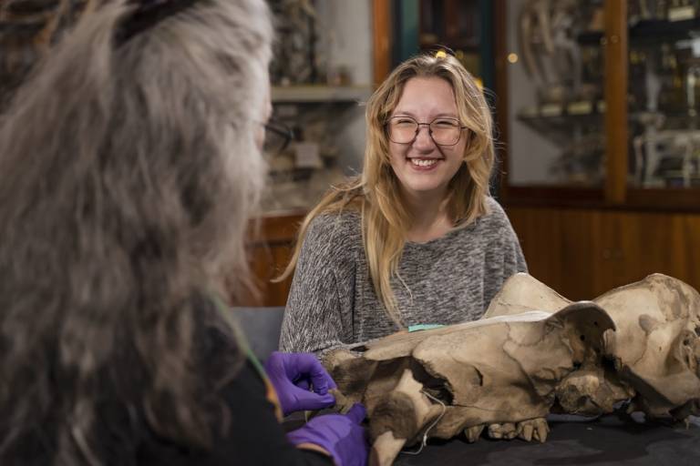 A young woman with long hair sits at a table in the Grant Museum in front of a large animal skull specimen, smiling towards an older woman who is facing away from the camera