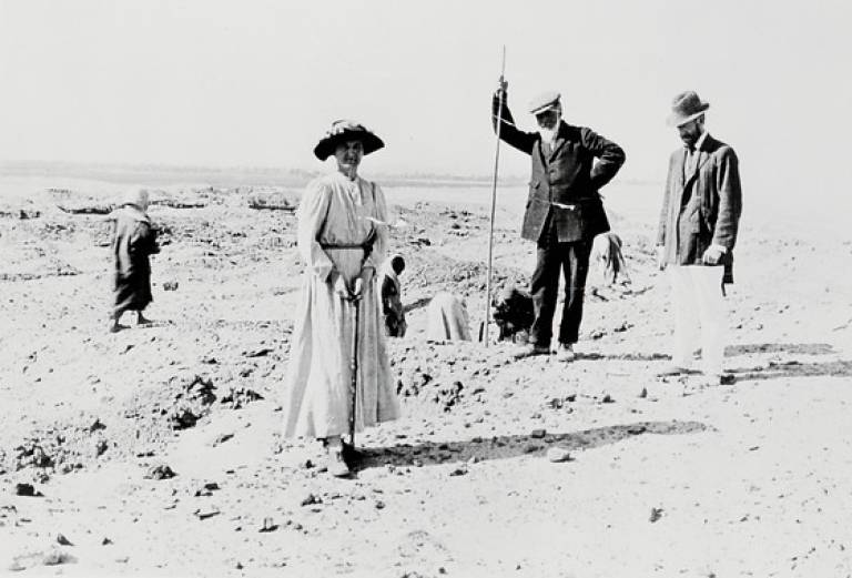 Black and white photograph of a group of people on a rocky, excavation site. A woman stands in the front left and a man to the right of her who is holding a long tool.