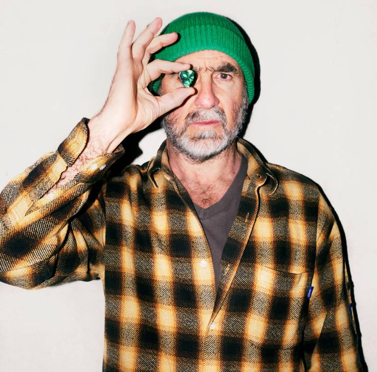 Photo of Eric Cantona looking directly at the camera. He is wearing a yellow & black check shirt and green beanie hat and holding up a green gem in front of his right eye