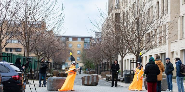 A group of people stand loosely together across a street on a housing estate, with two of them wearing long high-vis orange capes, and another filming them with a camera on a tripod