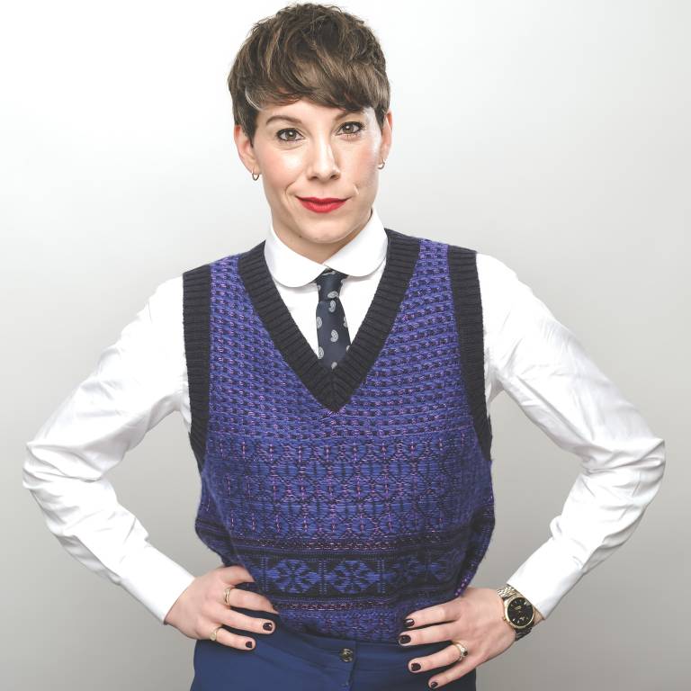 Photo of comedian Suzi Ruffell, standing with hands on hips and smiling wryly at the camera. She is wearing a white shirt and blue tie and a blue tank top
