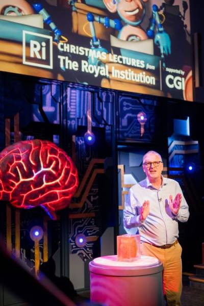 Photograph of a man standing on a lit stage with a brain specimen on a table in front of him. In the background there is a screen with text on reading 'Christmas Lectures from The Royal Institution'.