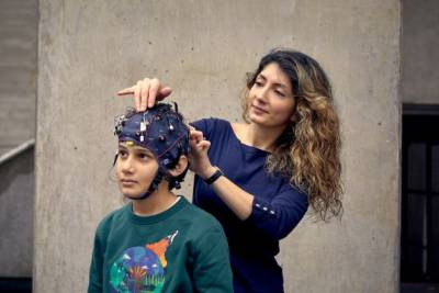 Bani Anvari stands behind a young boy who is wearing a brain-monitoring headset and adjusts the wiring