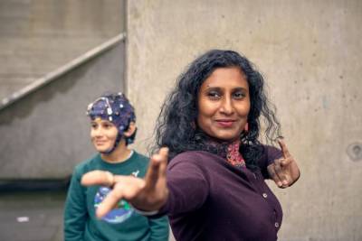 Suba Subramaniam reaches towards the camera as if dancing. A young boy wearing a brain-monitoring headset can be seen behind her.