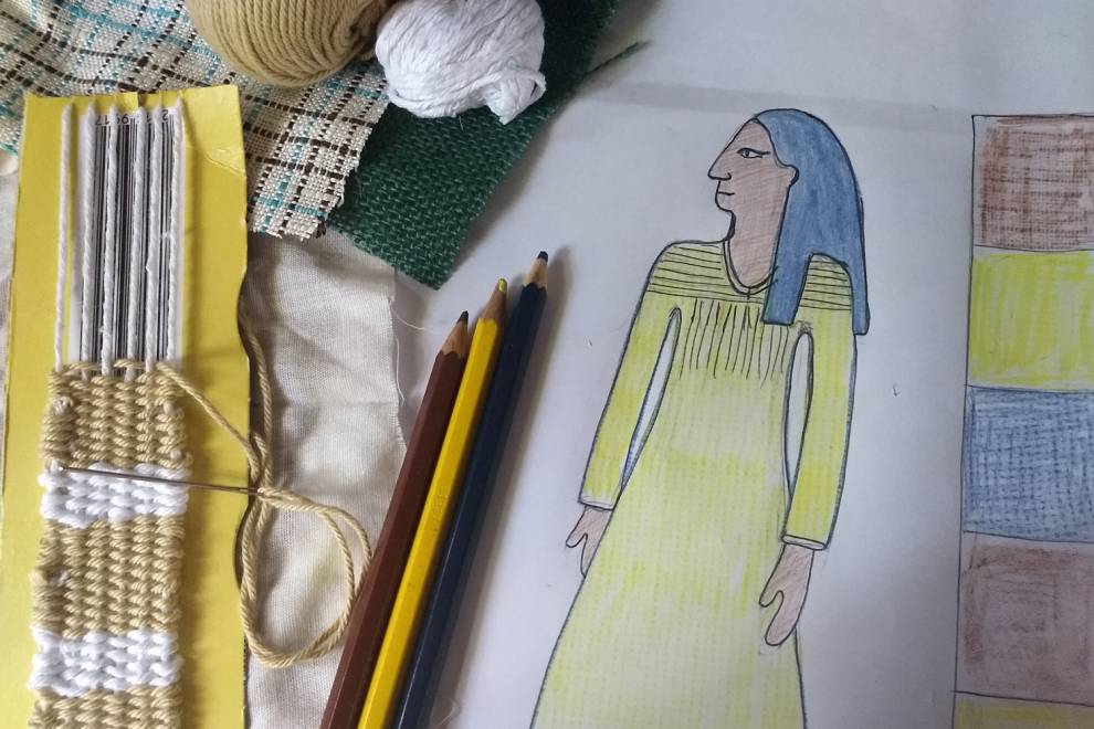 A close-up of a threaded homemade cardboard loom, some balls of yarn, coloured pencils and a drawing of a simplified ancient Egyptian figure