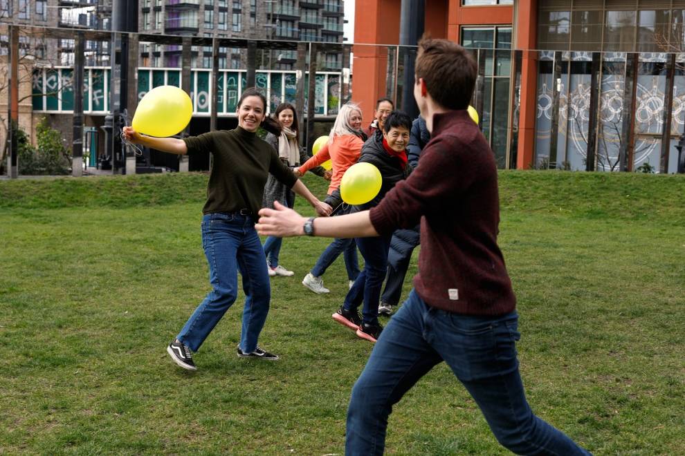 A group of smiling people run across an area of grass holding yellow balloons