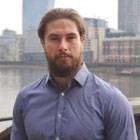 A young man with combed-back hair and a beard standing in front of an urban waterfront, wearing a blue shirt