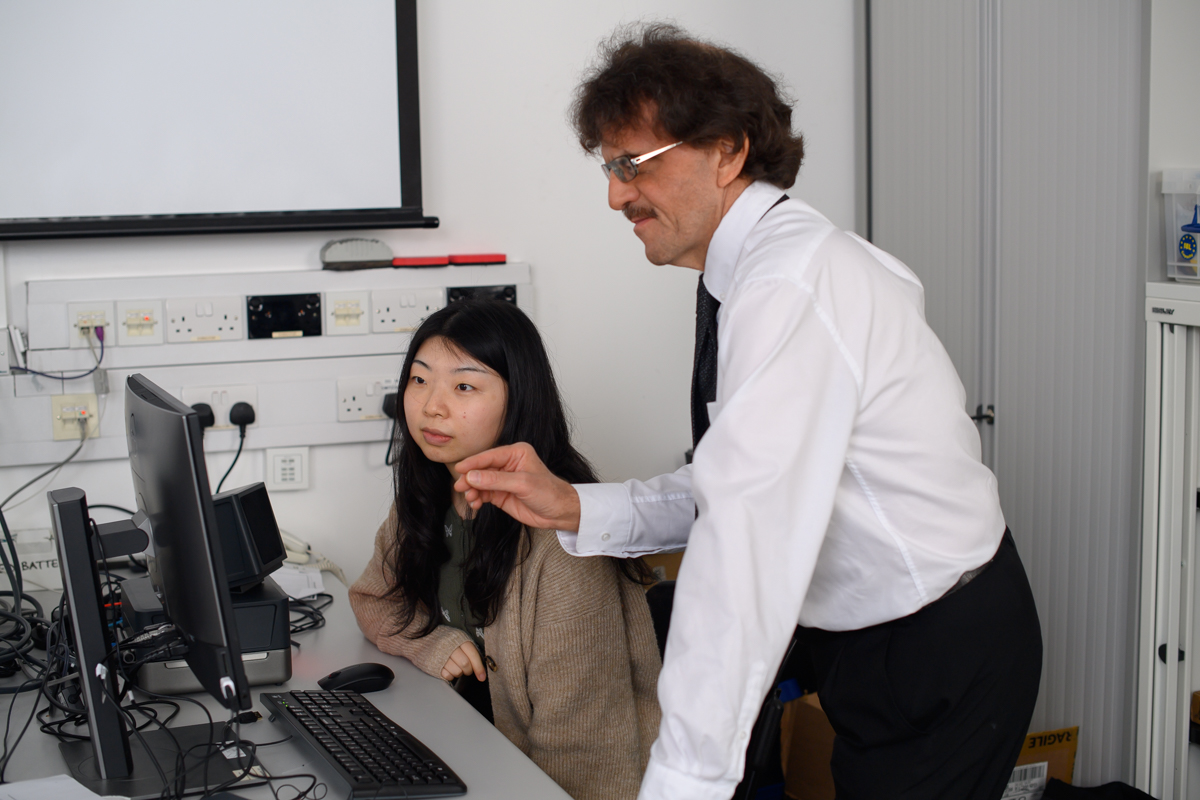 An image of a male teacher talking to a female student at a computer