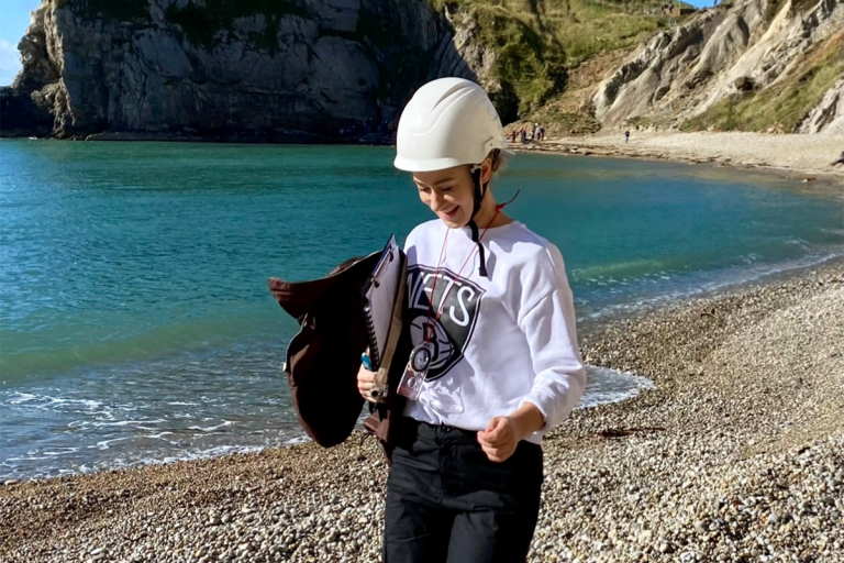 Amy Perrio wearing a white helmet and clipboard looking down whilst walking along a beach