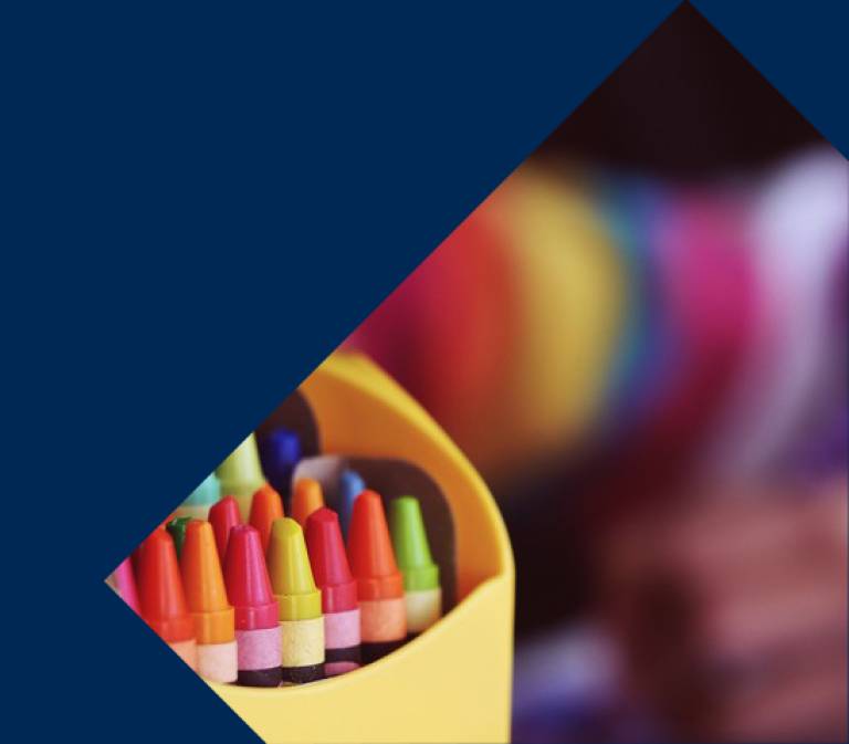 a brightly coloured pencil pot filled with colouring pencils is in the foreground. In the background is a child in a brightly coloured stripy top, colouring. The image is seen through a triangular cut out of a plain dark blue background. 