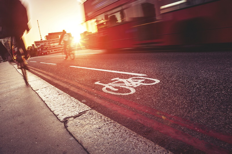 An image of two cyclists in a bicycle lane next to a bus, in front of a setting sun behind