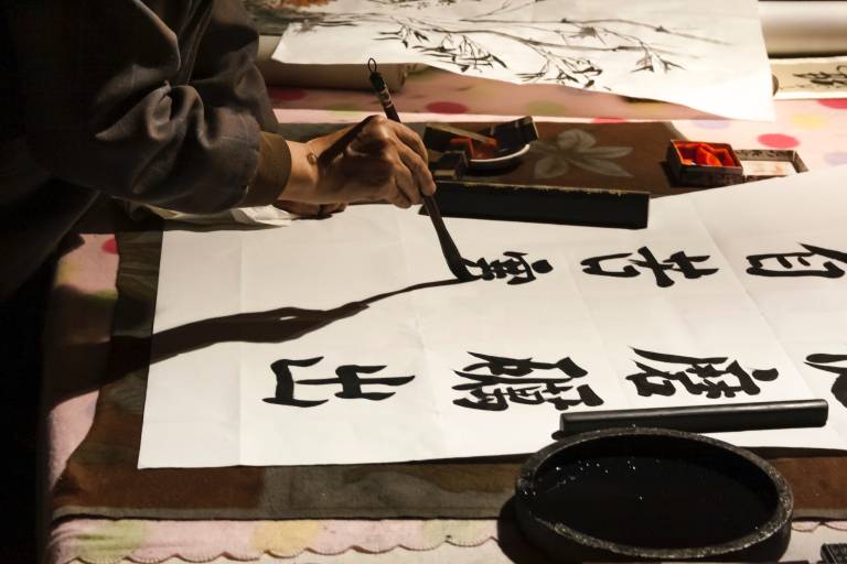 Image shows a person painting calligraphy with a brush, paper and ink.