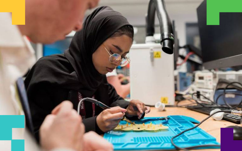 A student in a headscarf solders an electronic circuit with a soldering iron.