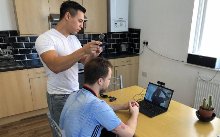 One man cuts another's hair, sitting at a kitchen table, while being guided by a professional barber via a video call