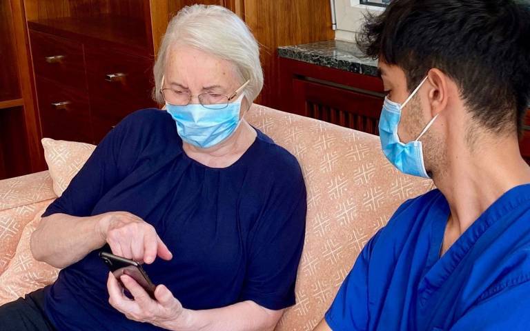 A medical professional sits on a sofa with an older person who is looking at a mobile phone