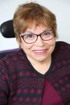A headshot of Judy Heumann, a cis-gender white woman who is a wheelchair user with short brown hair. She is wearing red glasses and a maroon and black embroidered sweater with the top buttoned with a matching maroon shirt underneath. She is smiling kindly