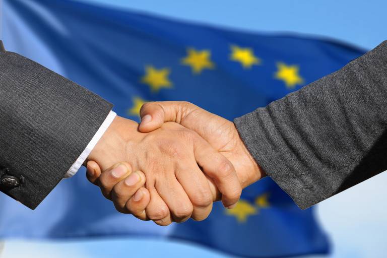 A close-up photo of a handshake between two people with the EU flag in the background