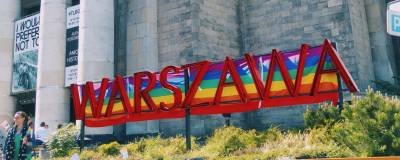 Photo of a large red sign reading WARSZAWA, with Gay Pride flags hung behind it to give the appearance of a rainbow striped background.
