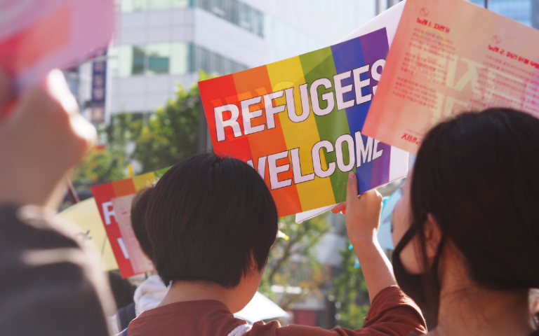 a protest with someone holding a sign with the LGBTQ+ colours saying refugees welcome