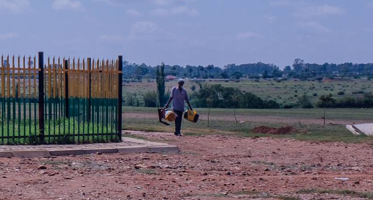 Image of a black person carrying two large items in either hand while walking in an open space.