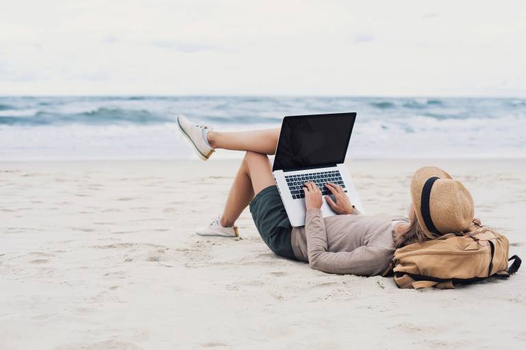 Woman uses laptop whilst lying on a beach. 