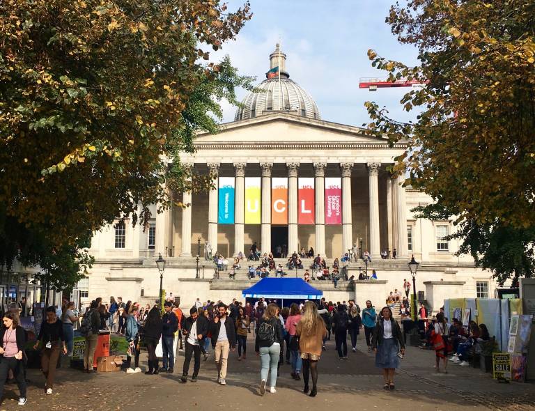 A picture of the UCL campus with multi coloured banners spelling out UCL. There are some trees and students on campus.