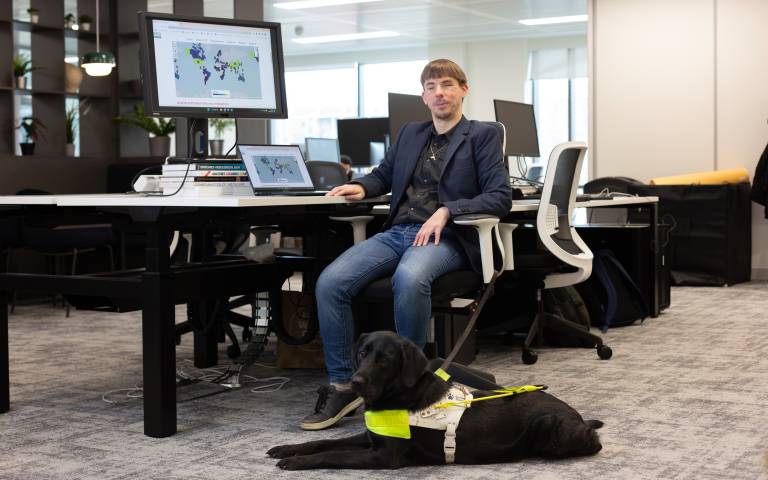 Dr Daniel Hajas sitting at a computer workspace with his guide dog.