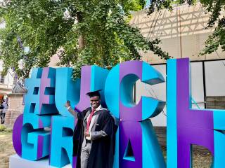 Student Patrick, standing in a graduation gown next to the UCL sign smiling